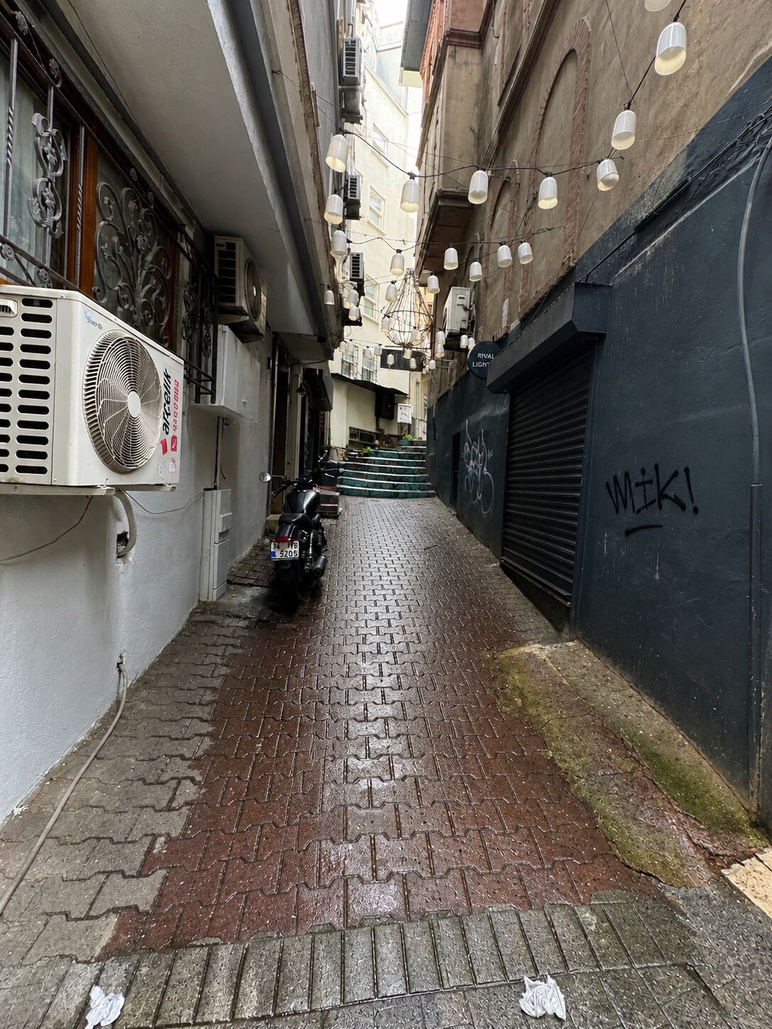 A narrow alleyway with a parked motorcycle, a fan on the left, and a closed shutter on the right.