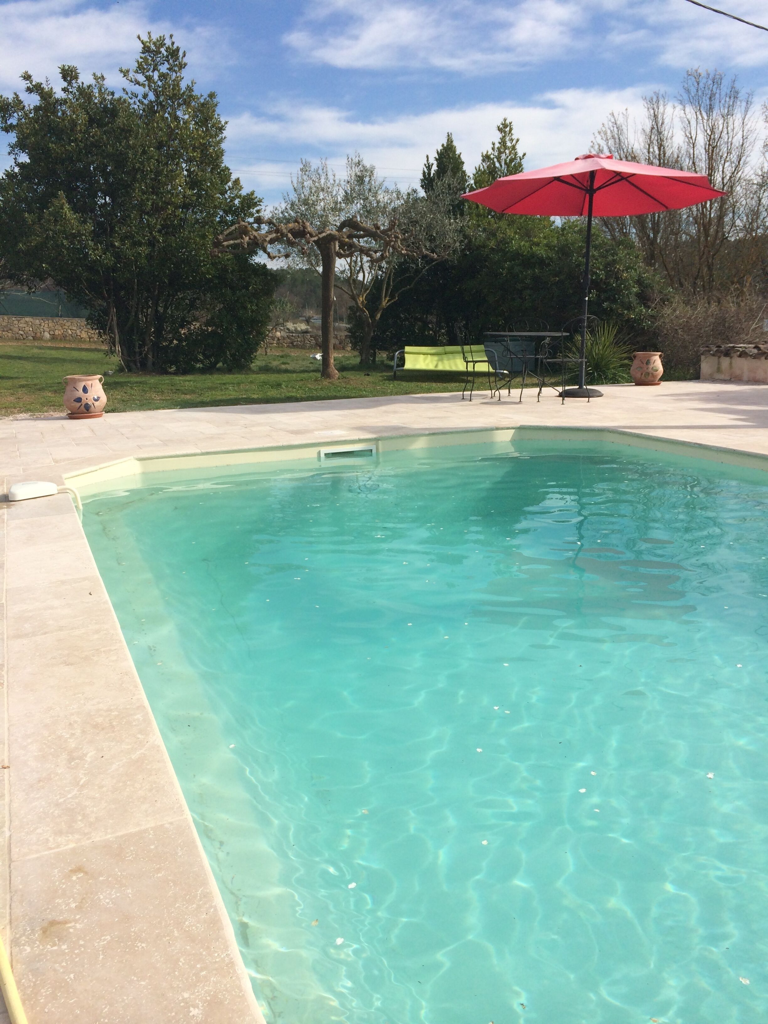 A clear swimming pool with a tiled edge, surrounded by a patio area with a red umbrella, outdoor furniture, and potted plants.