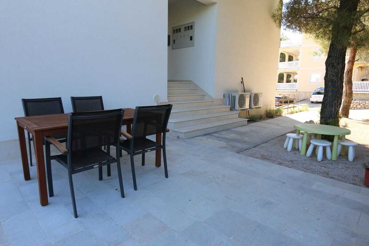 Une terrasse avec une table et des chaises en bois, une petite table ronde avec des tabourets, un escalier et un bâtiment en arrière-plan.