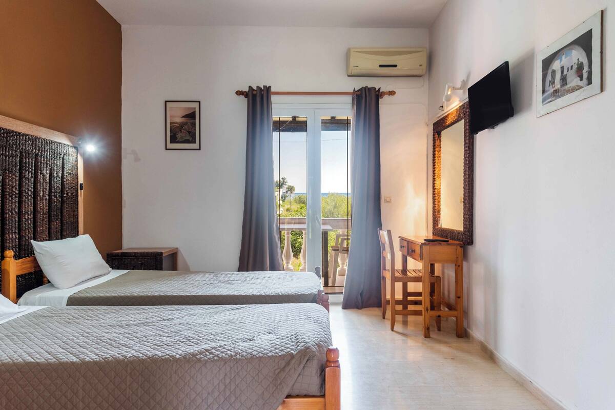 A hotel room with two beds, a desk, a chair, a TV, and a balcony with curtains.