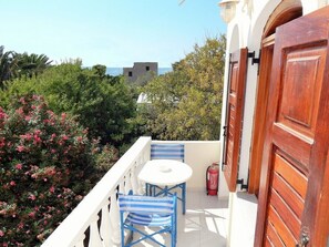 A balcony with a white table and blue chairs, surrounded by lush greenery and a view of the sea. - Evelina Pension (Santorini)