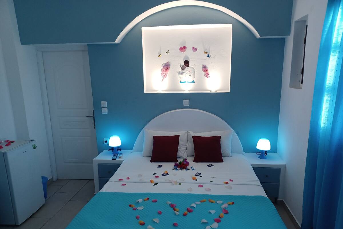 Una habitació d'hotel amb un llit, dues làmpades de nit, una tauleta de nit, un televisor fixat a la paret i una decoració amb forma de cor al llit.