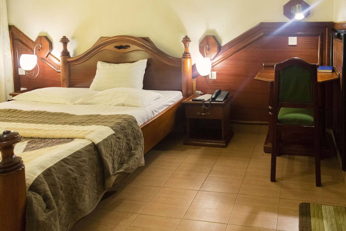 Habitación de hotel con cama de madera, mesita de noche con lámpara, una silla y escritorio con silla.