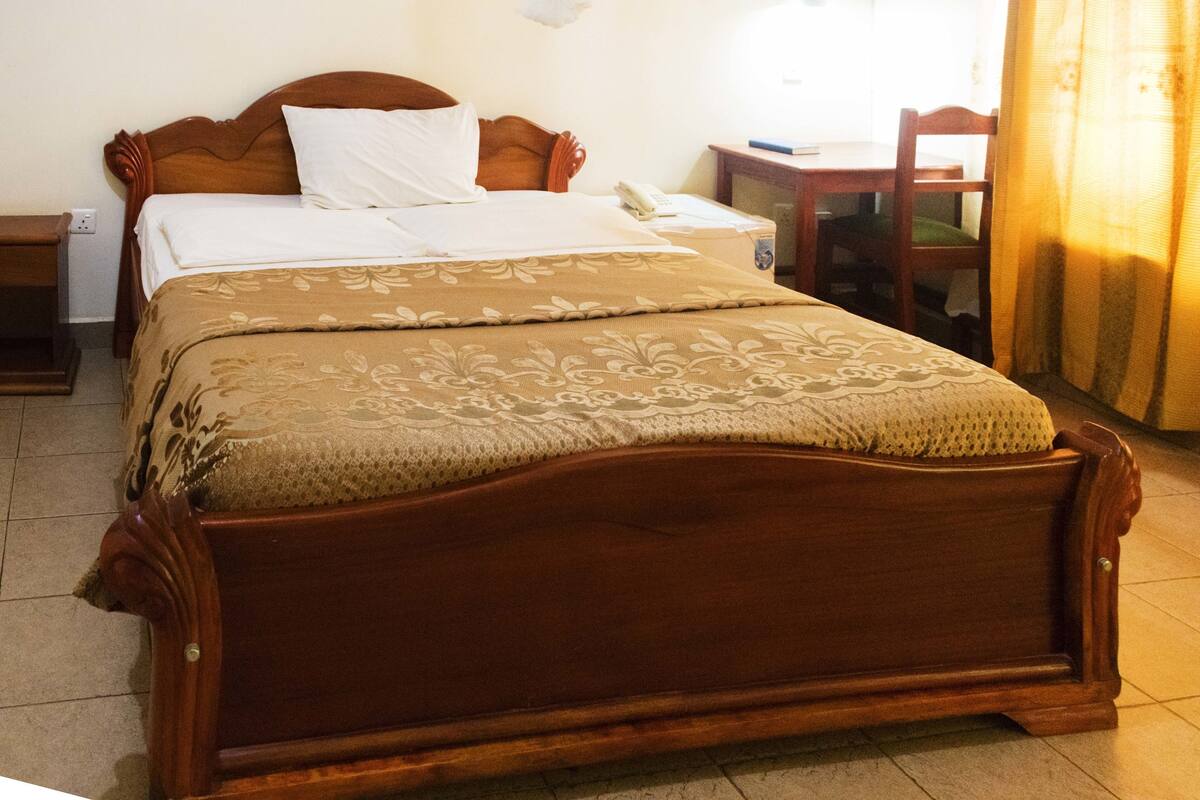 Una cama de madera con colcha estampada, una mesita de noche, una silla y una pequeña mesa con un teléfono.