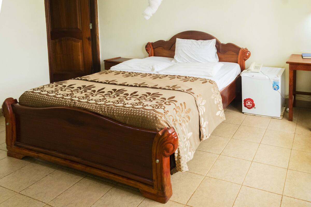 Una cama de madera con una colcha estampada, una mesita de noche con un teléfono y un refrigerador pequeño.