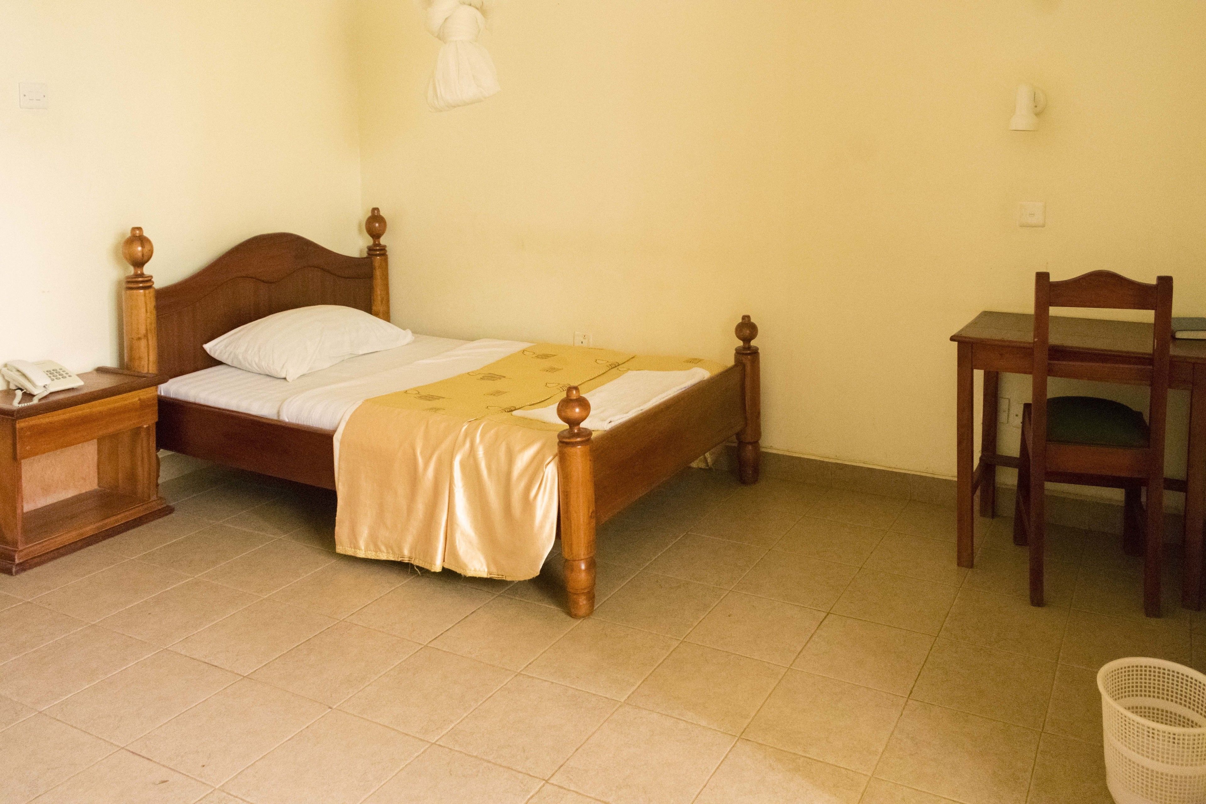 Una cama individual con cabecera de madera, una mesita de noche con teléfono, una silla y una pequeña mesa con lámpara.