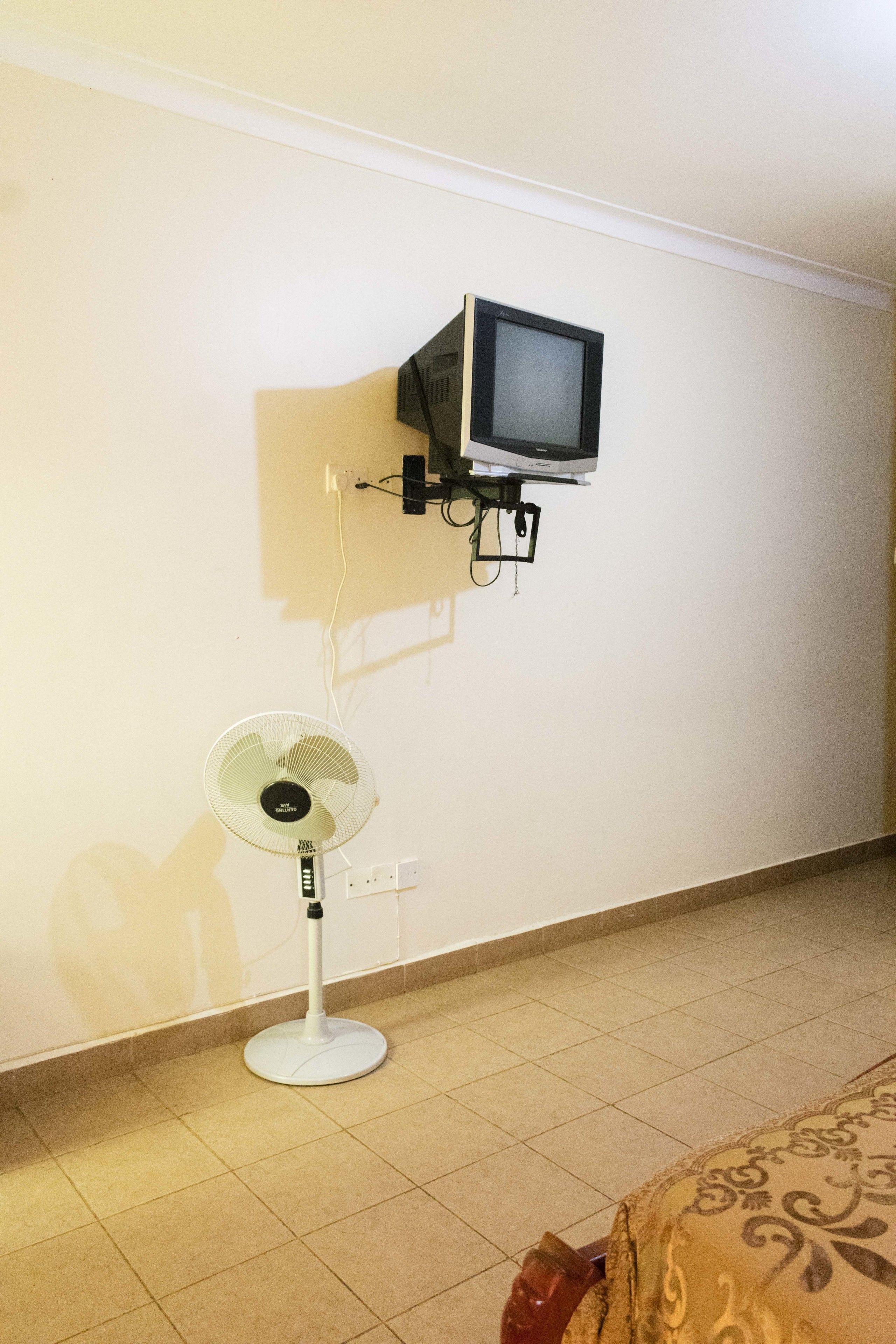 A room with a bed, a standing fan, and a mounted television on the wall.