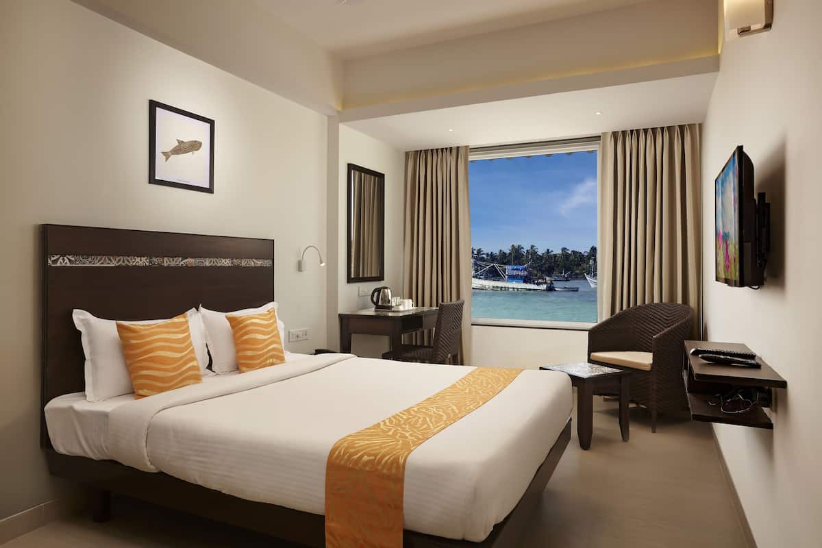 A hotel room with a bed, a desk, a chair, a TV, and a window with a view of a body of water and palm trees.