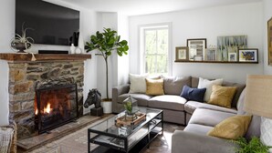 A living room with a stone fireplace, a grey sofa, a glass coffee table, and a large window. - 1805 Farmhouse with Heated Pool in Harpers Ferry (Harpers Ferry)