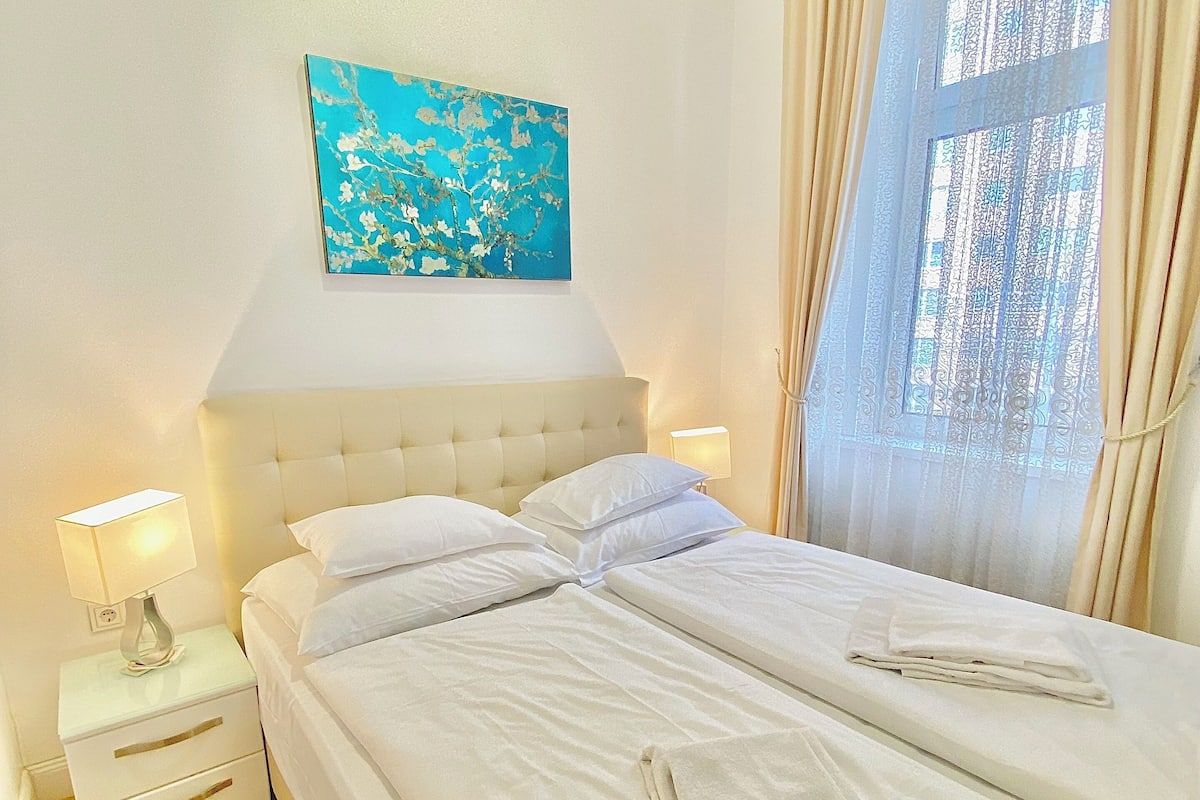 A neatly made bed with white linens and towels, a bedside lamp, a small white nightstand, a wall-mounted painting, and a window with curtains.