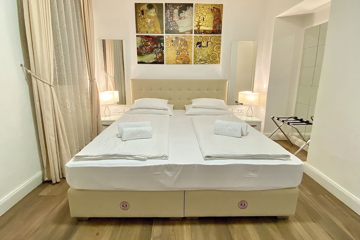 A double bed with two pillows, a headboard, and a bedside table with a lamp. There is a window with curtains, a wall with a multi-panel artwork, and a door with a towel rack.