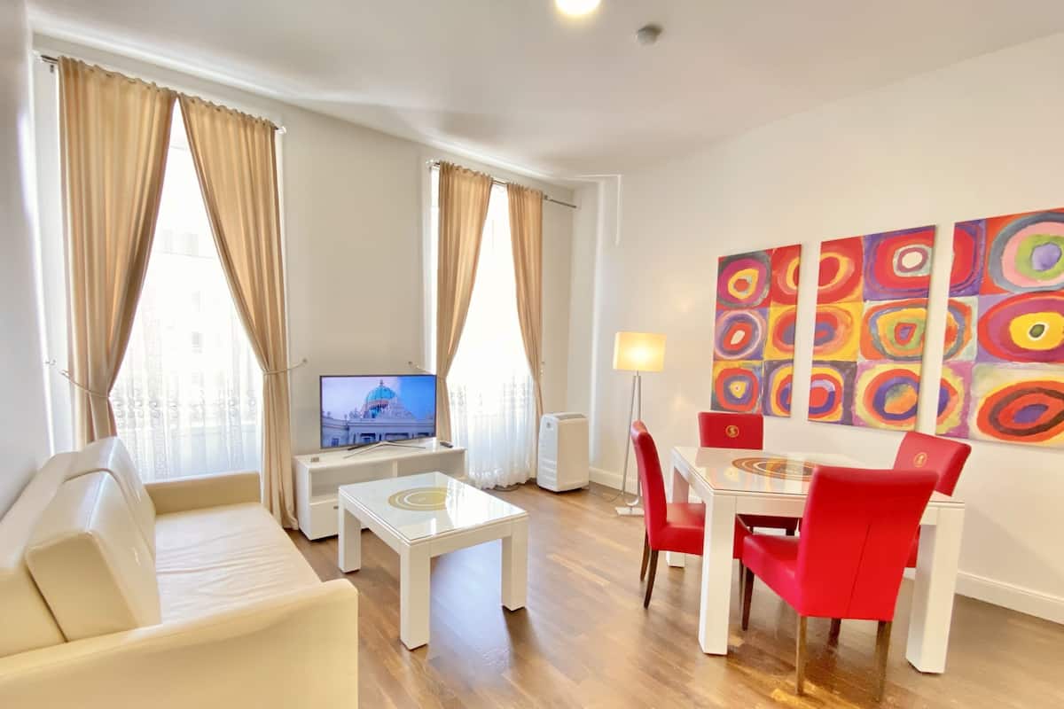 A modern living room with a sofa, a small table, a TV, and a dining area with red chairs.