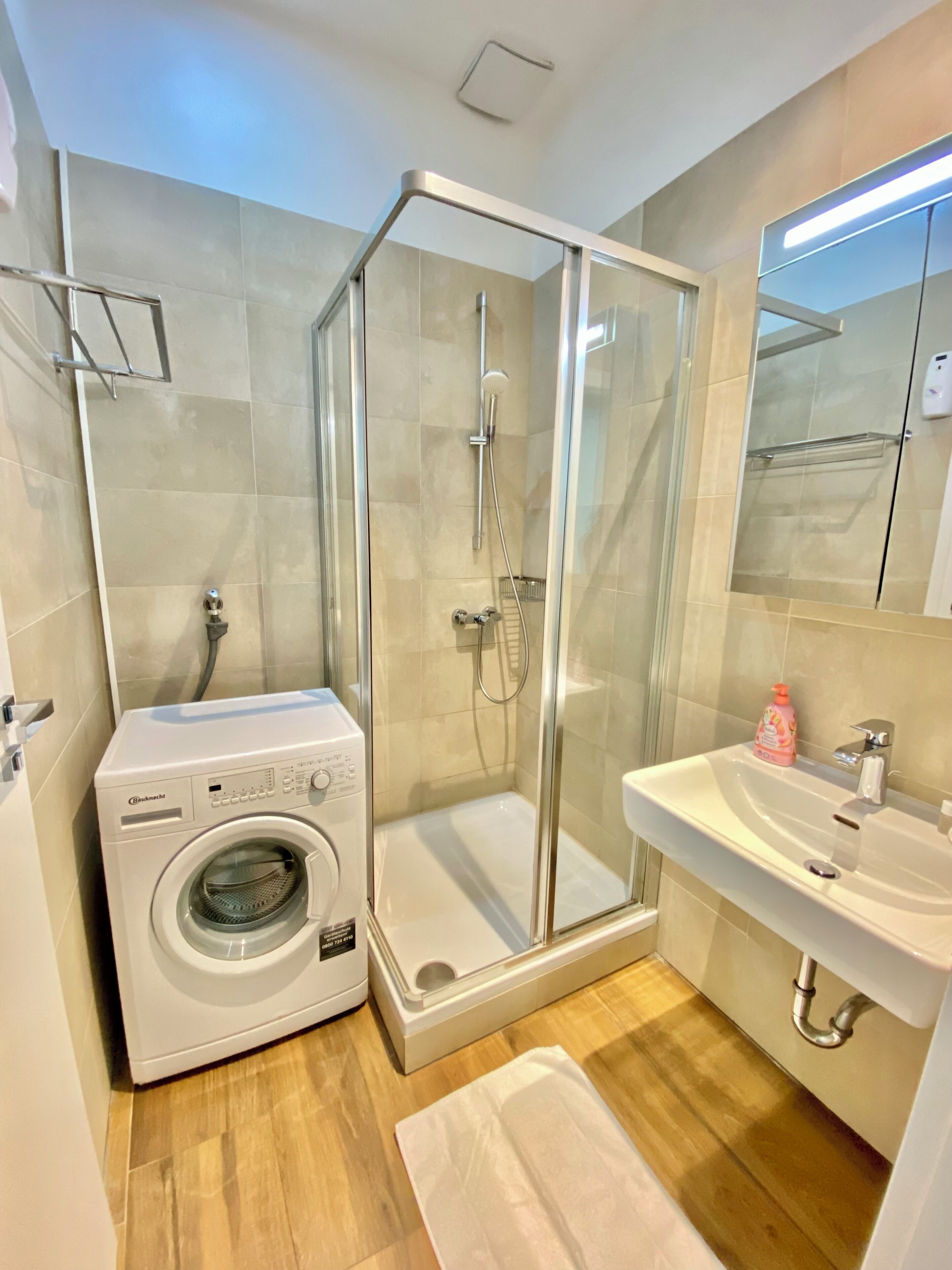 A modern bathroom with a glass-enclosed shower, a built-in washing machine, and a sink with a mirror above it.