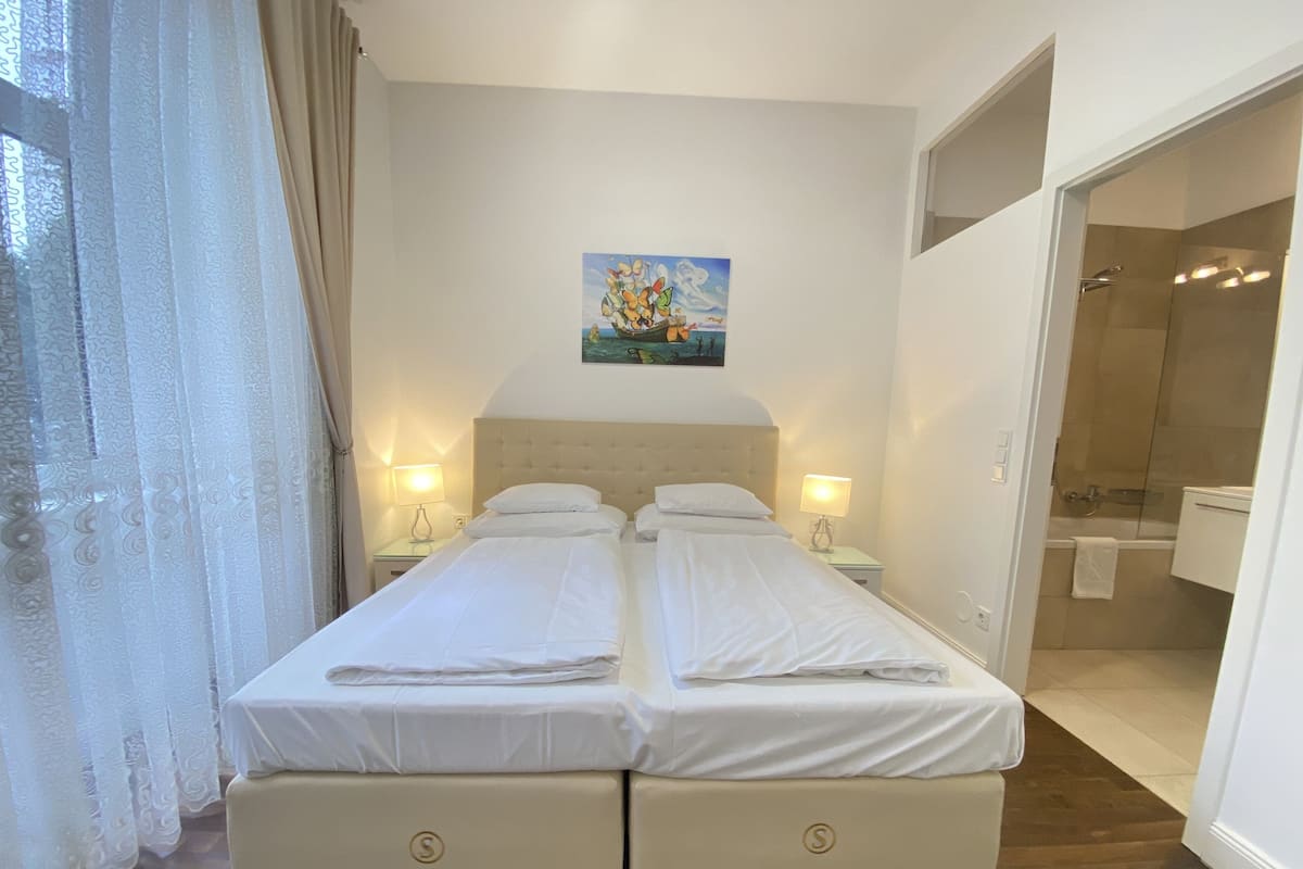 A double bed with two nightstands, a painting above the bed, and a bathroom visible through an open door.