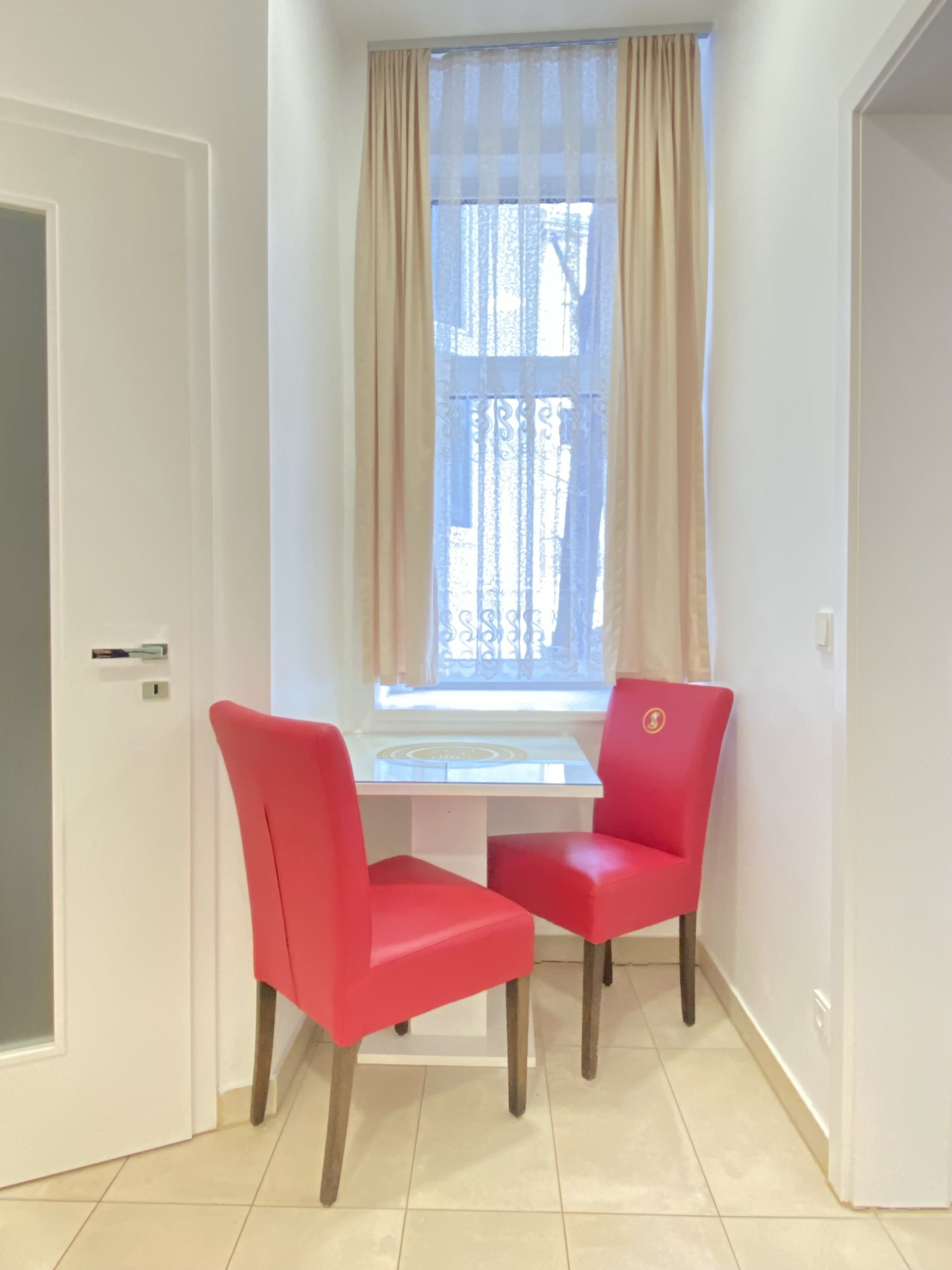 A room with a white desk and two red chairs, a window with beige curtains, and a tiled floor.