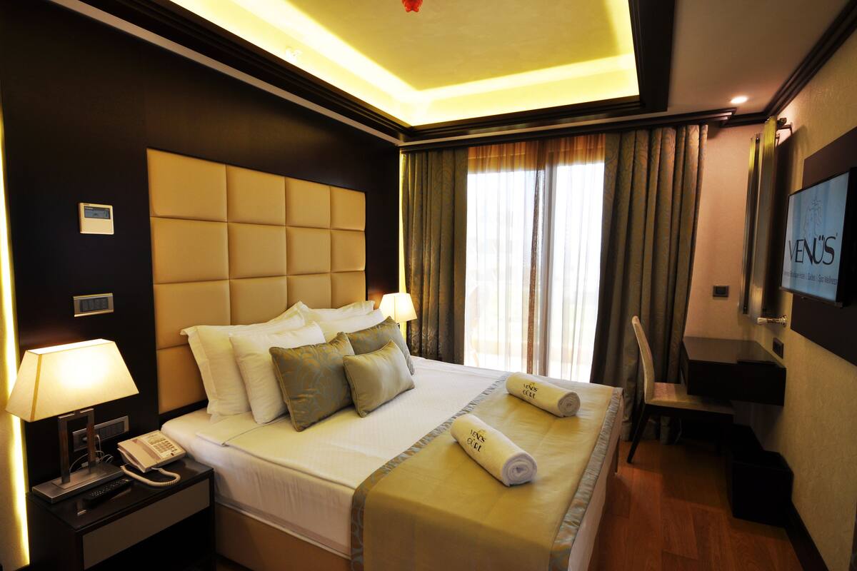 A hotel room with a large bed, a bedside table with a lamp and phone, a TV on the wall, and a desk with a chair.