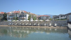 Ein Strandhotel mit mehreren Balkonen, einer Steinmauer und einer Reihe von Liegestühlen.