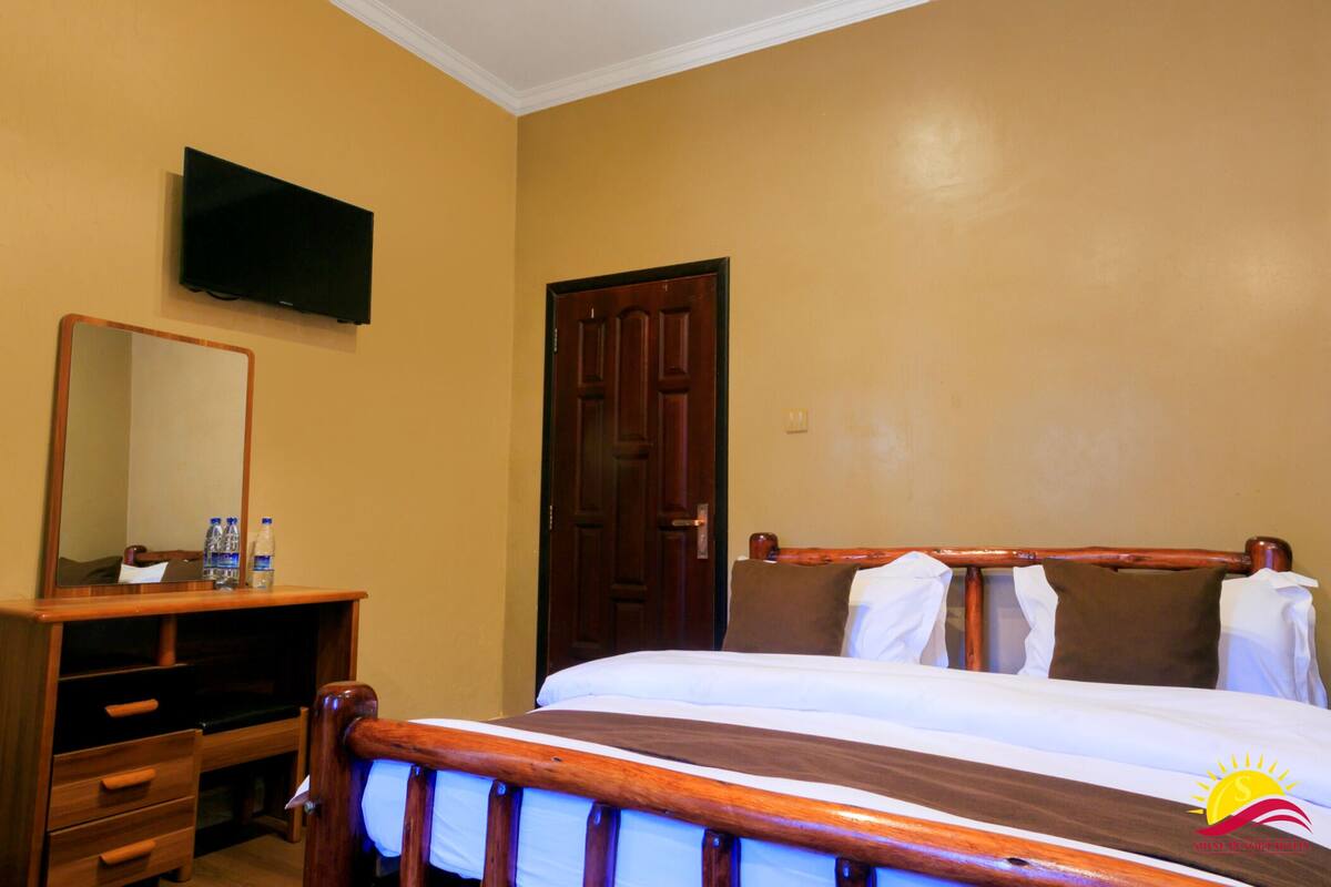 A hotel room with a wooden bed, a wooden dresser with a mirror, a flat-screen TV mounted on the wall, and a door.