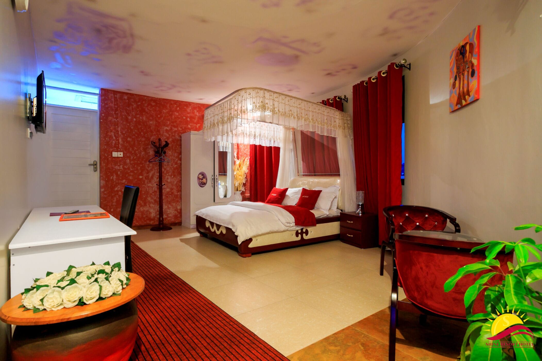 A bedroom with a four-poster bed, a desk with a vase of flowers, a red chair, and a large window with curtains.