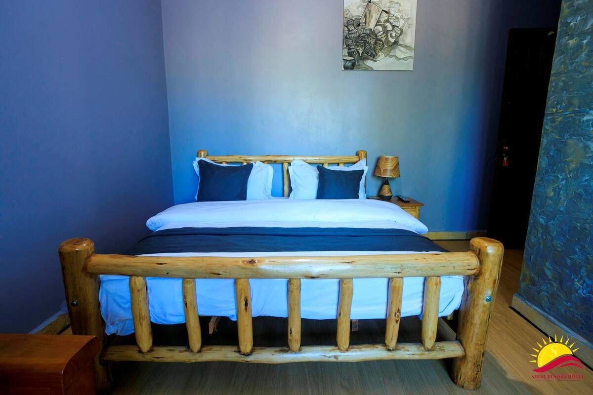 A wooden-framed bed with a white and blue bedding set, a nightstand with a lamp, and a blue wall with a framed artwork.