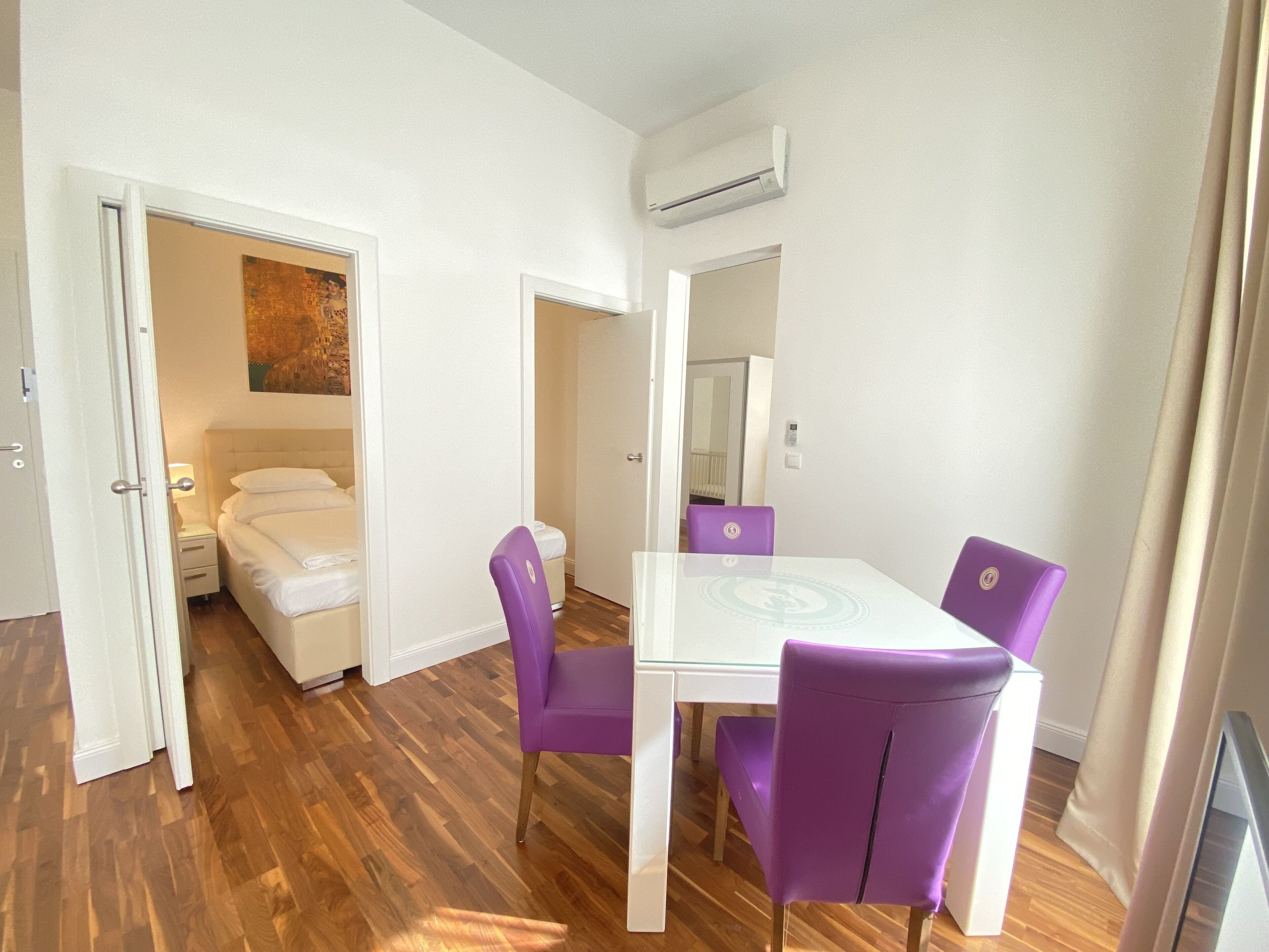 A room with a white bed, a white table with purple chairs, and a white air conditioning unit on the wall.