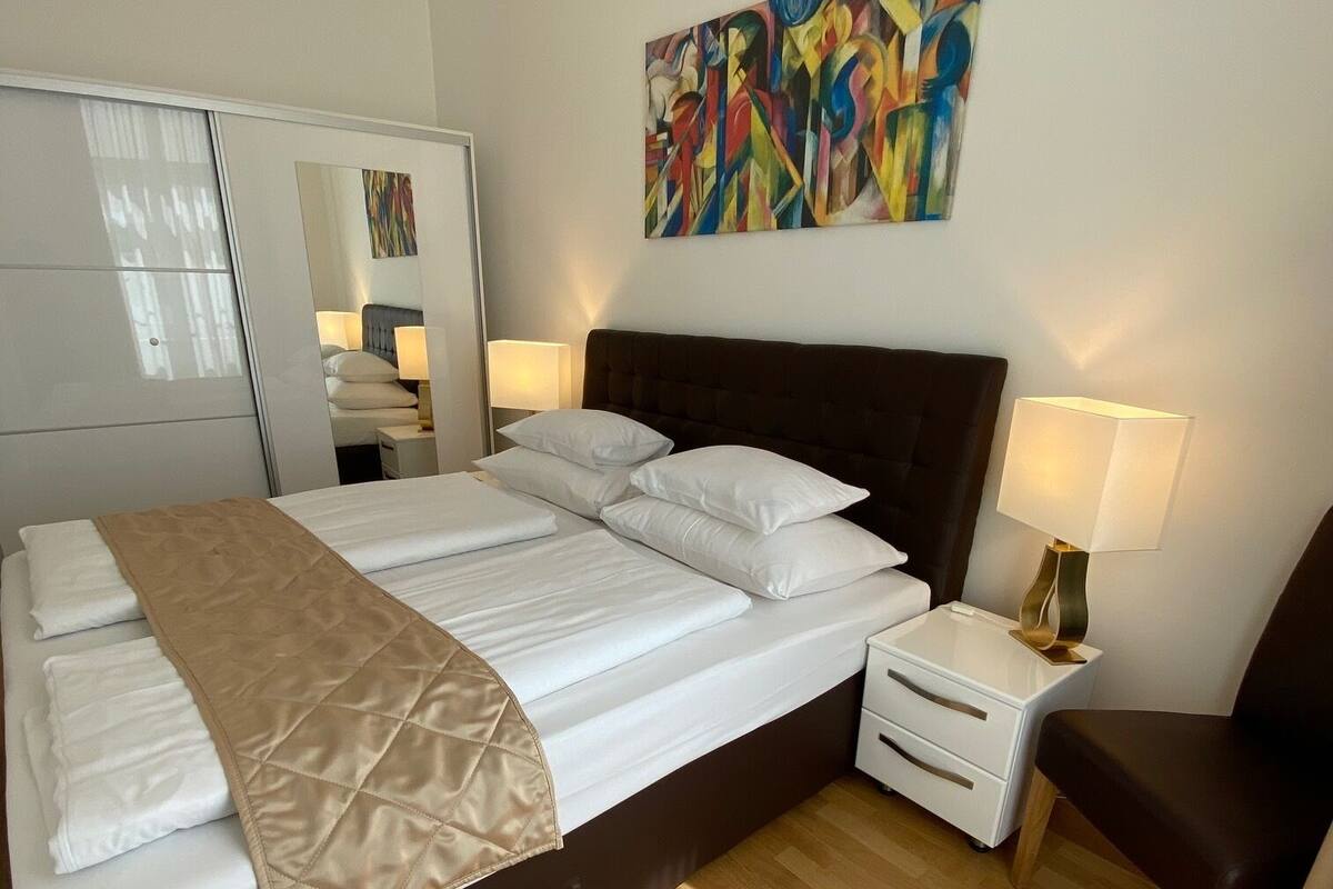 Ein modernes Schlafzimmer mit einem großen Bett, Nachttischen, einem Sessel und einem farbenfrohen, abstrakten Gemälde an der Wand.