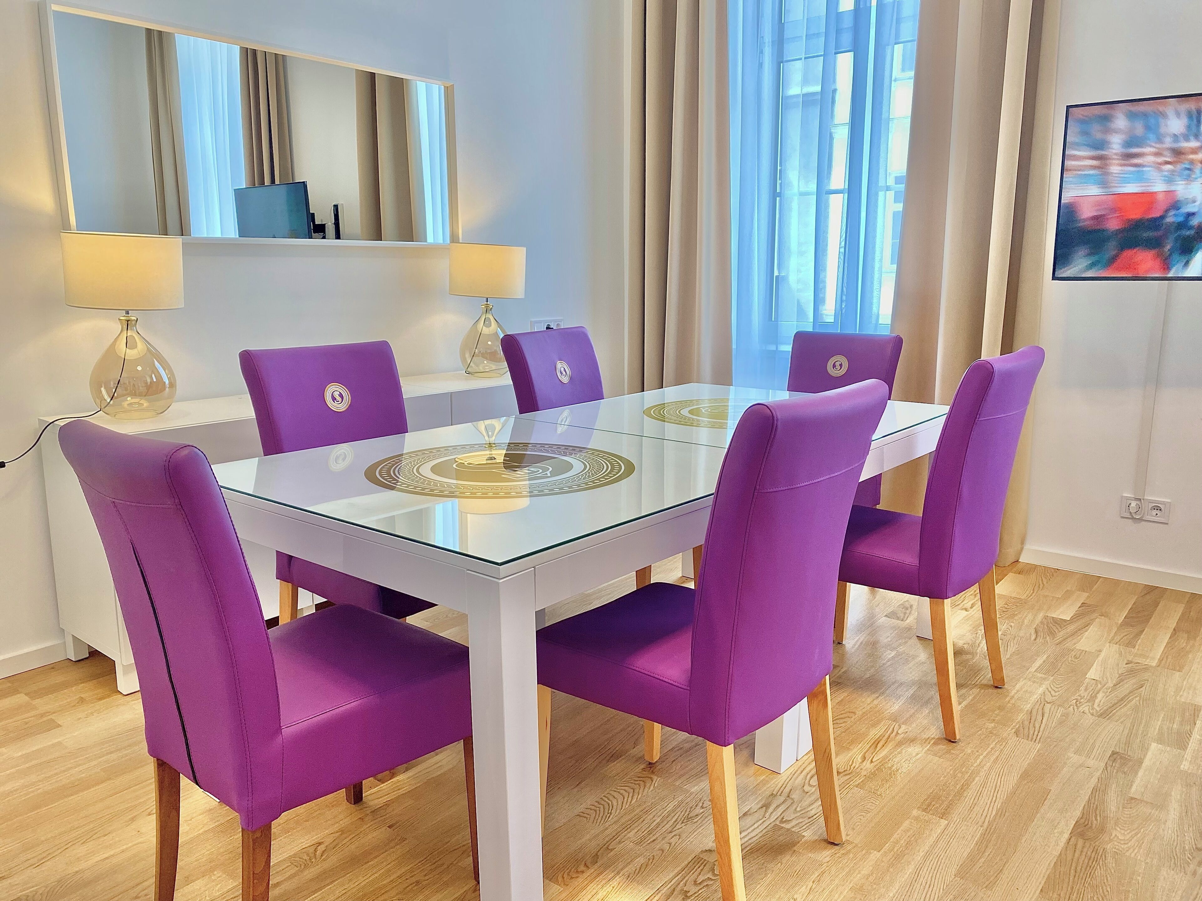 A dining area with a glass-top table, purple chairs, a lamp, and a painting on the wall.