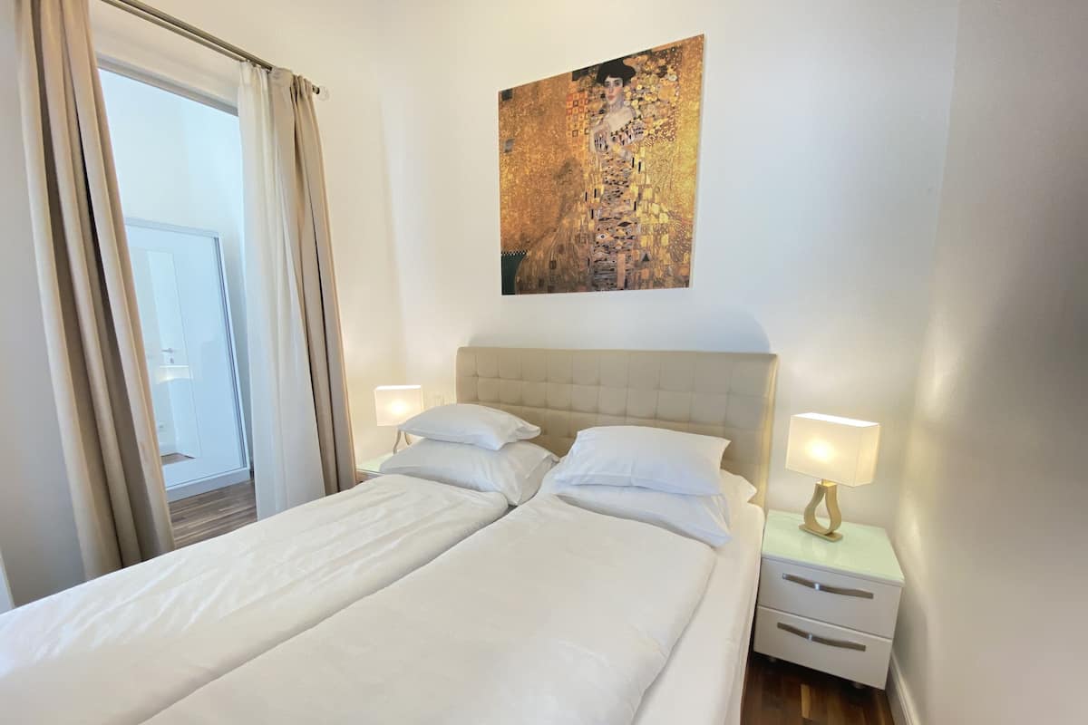 Ein Hotelzimmer mit Doppelbett, zwei Nachttischen mit Lampen, einem Bild an der Wand und Blick durch ein mit Vorhängen drapiertes Fenster.