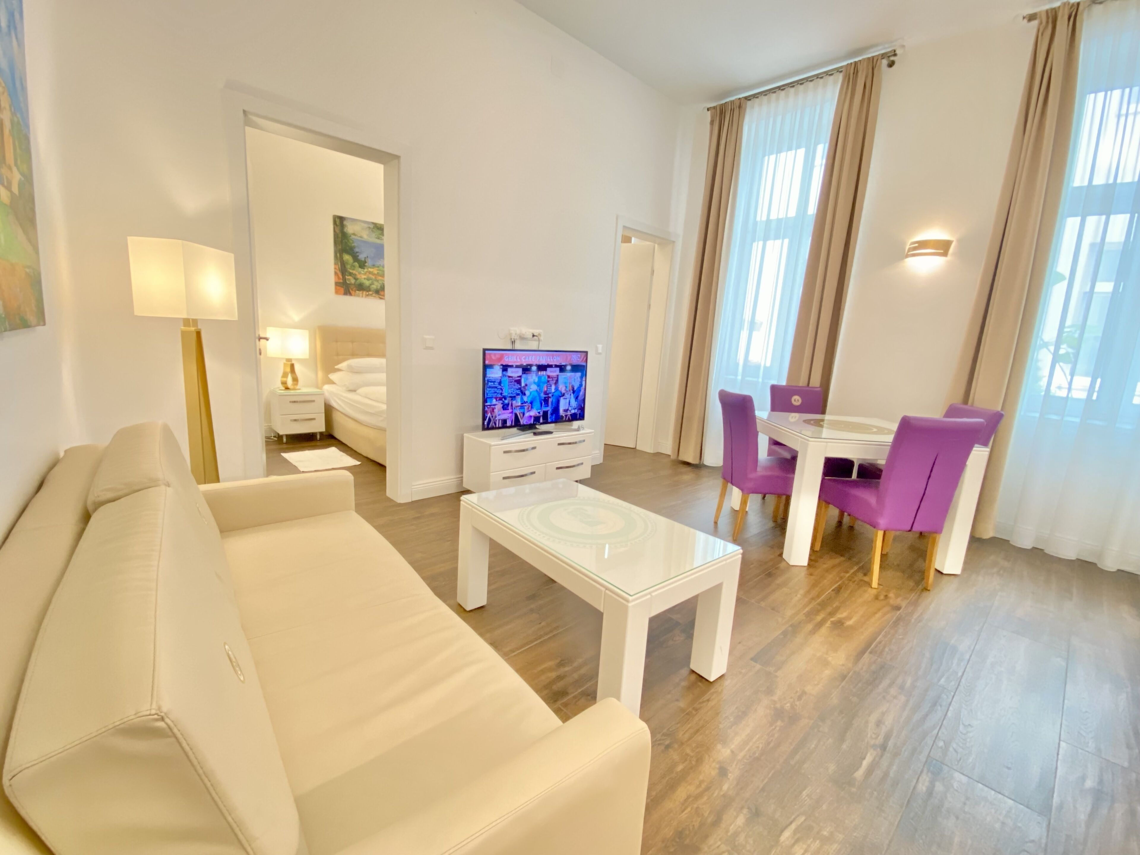 Ein modernes Hotelzimmer mit einer Couch, einem Couchtisch, einem Fernseher, einem Esstisch mit violetten Stühlen und einem großen Fenster mit Vorhängen.