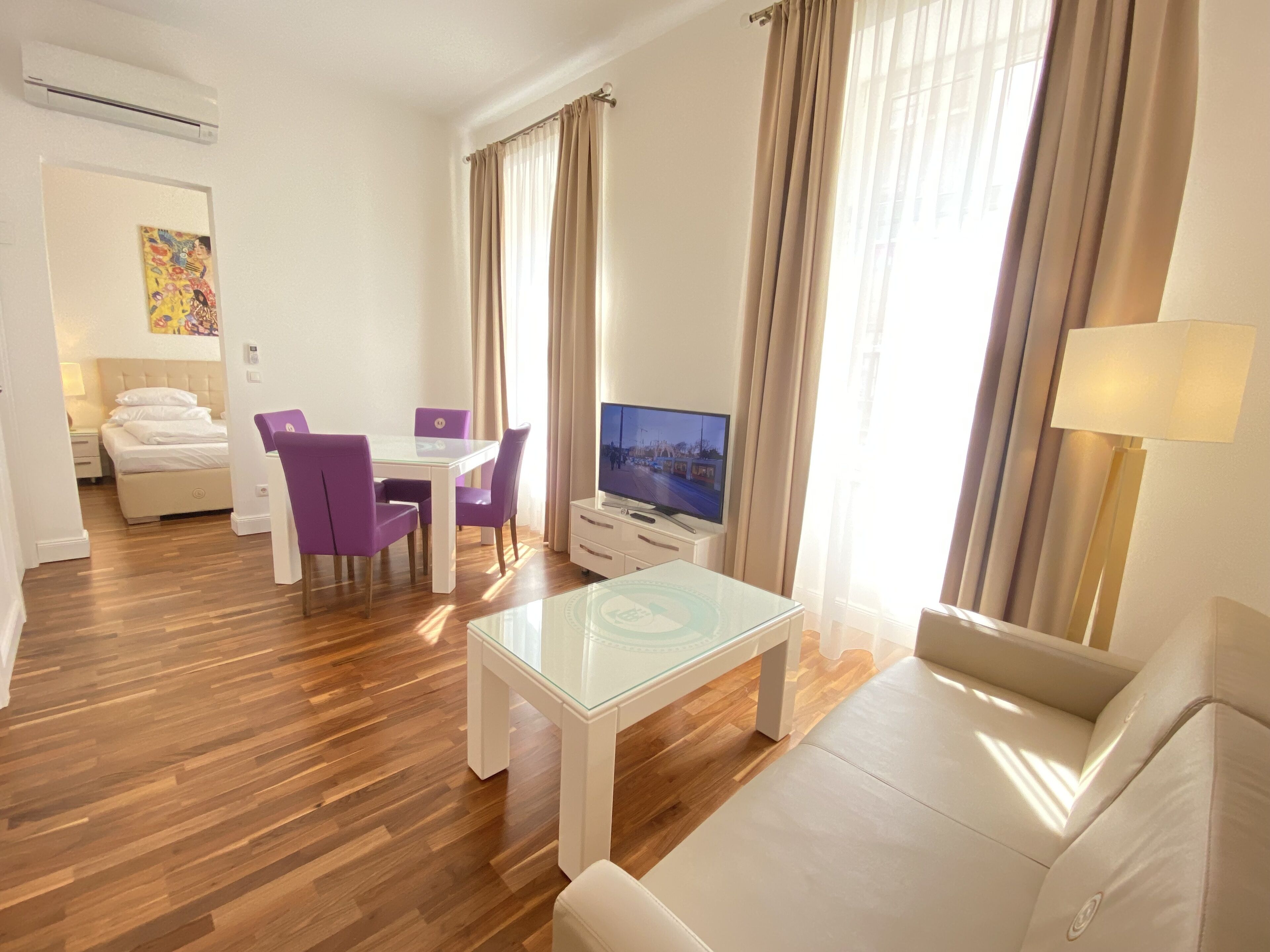A modern hotel room with a wooden floor, a glass coffee table, a sofa, a TV, and a dining area with purple chairs.