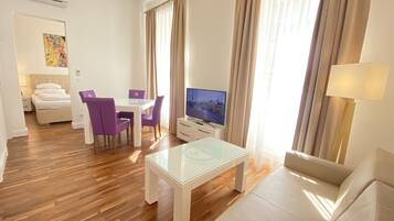 Une chambre d’hôtel moderne avec un plancher en bois, une table basse en verre, un canapé, une télévision et un coin repas avec des chaises violettes.