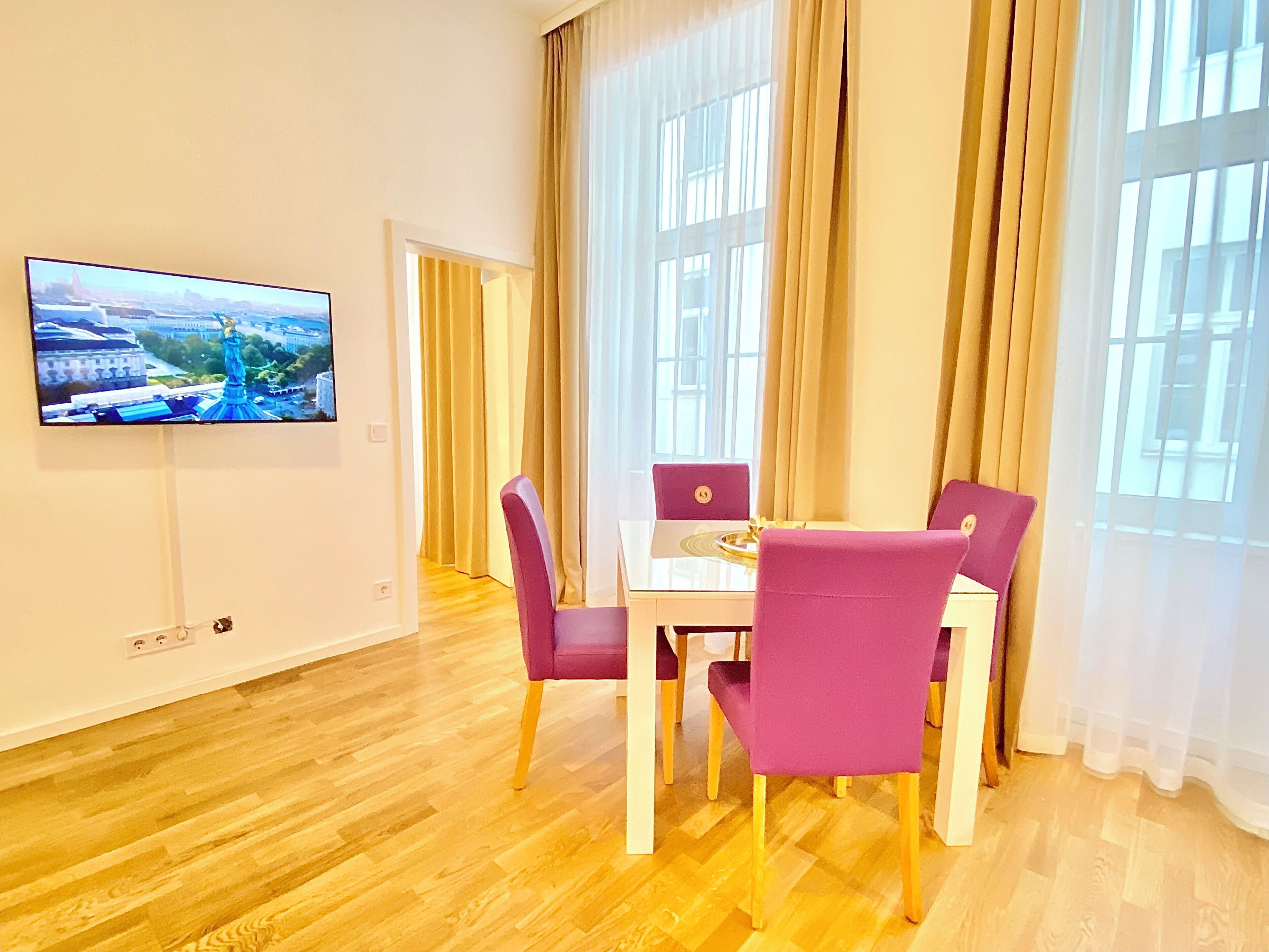 Un comedor moderno con piso de madera, una mesa de vidrio y sillas moradas. Un televisor de pantalla plana está montado en la pared.