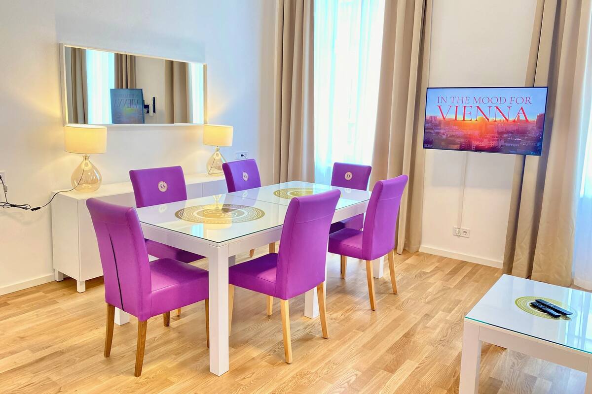 Ein moderner Essbereich mit Glastisch und violetten Stühlen, ein Fernseher zeigt 'IN THE MOOD FOR VIENNA', und eine weiße Konsole mit Lampen.