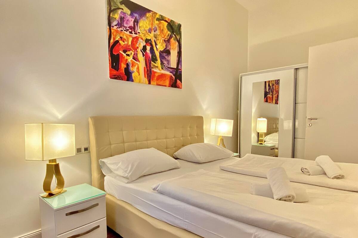 Ein Hotelzimmer mit einem großen Bett, zwei Nachttischen mit Lampen, einem Spiegel und einem farbenfrohen, abstrakten Gemälde an der Wand.