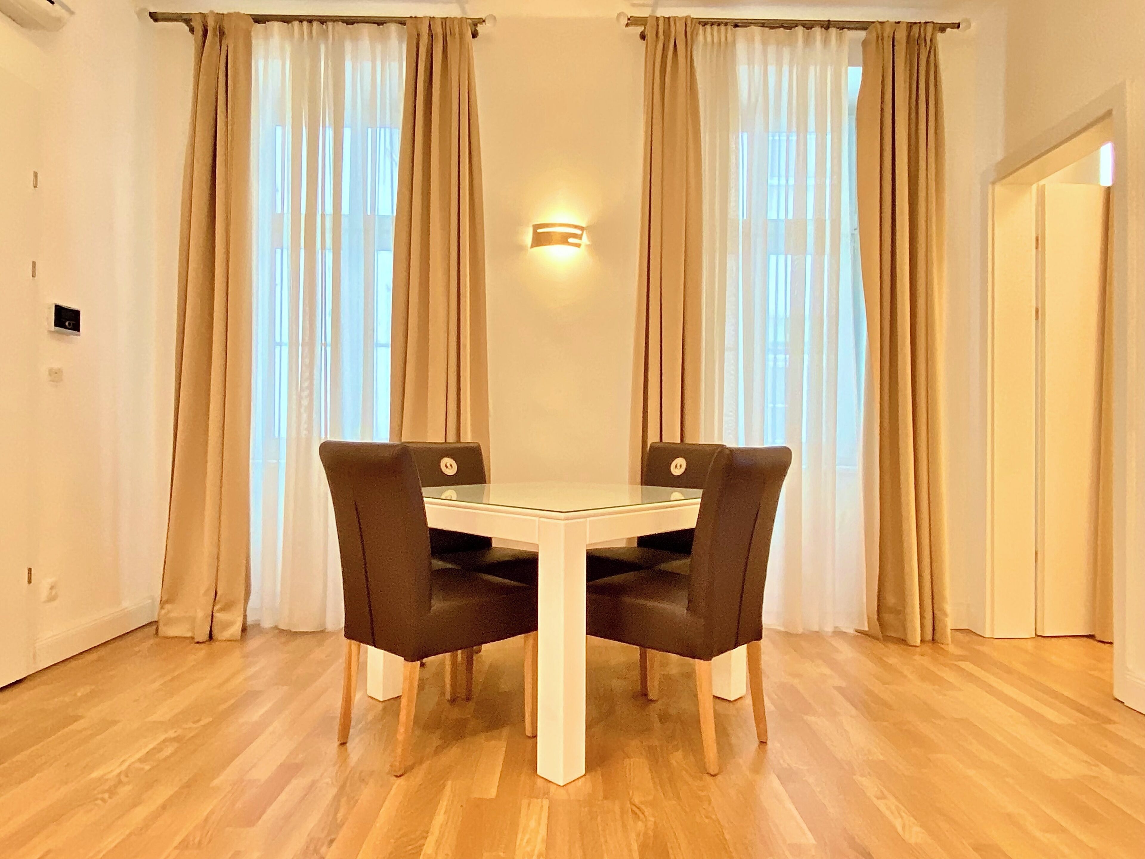 A dining area with a white table and four chairs, beige curtains, and a wall-mounted light fixture.