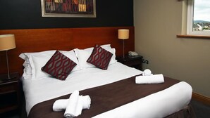 A neatly made bed with white linens, two patterned pillows, and two rolled towels on a brown bed runner. There is a nightstand with a lamp and a phone, and a painting on the wall. - Ashford Court Boutique Hotel (Ennis)