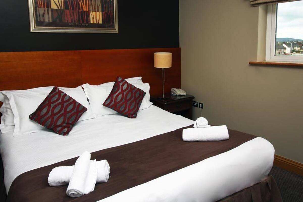 A neatly made bed with white linens, two patterned pillows, and two rolled towels on a brown bed runner. There is a nightstand with a lamp and a phone, and a painting on the wall.