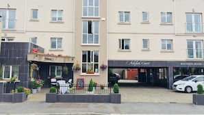 A hotel with a central entrance, a flower-adorned outdoor seating area, and a visible storefront named "Ashford Court." - Ashford Court Boutique Hotel (Ennis)