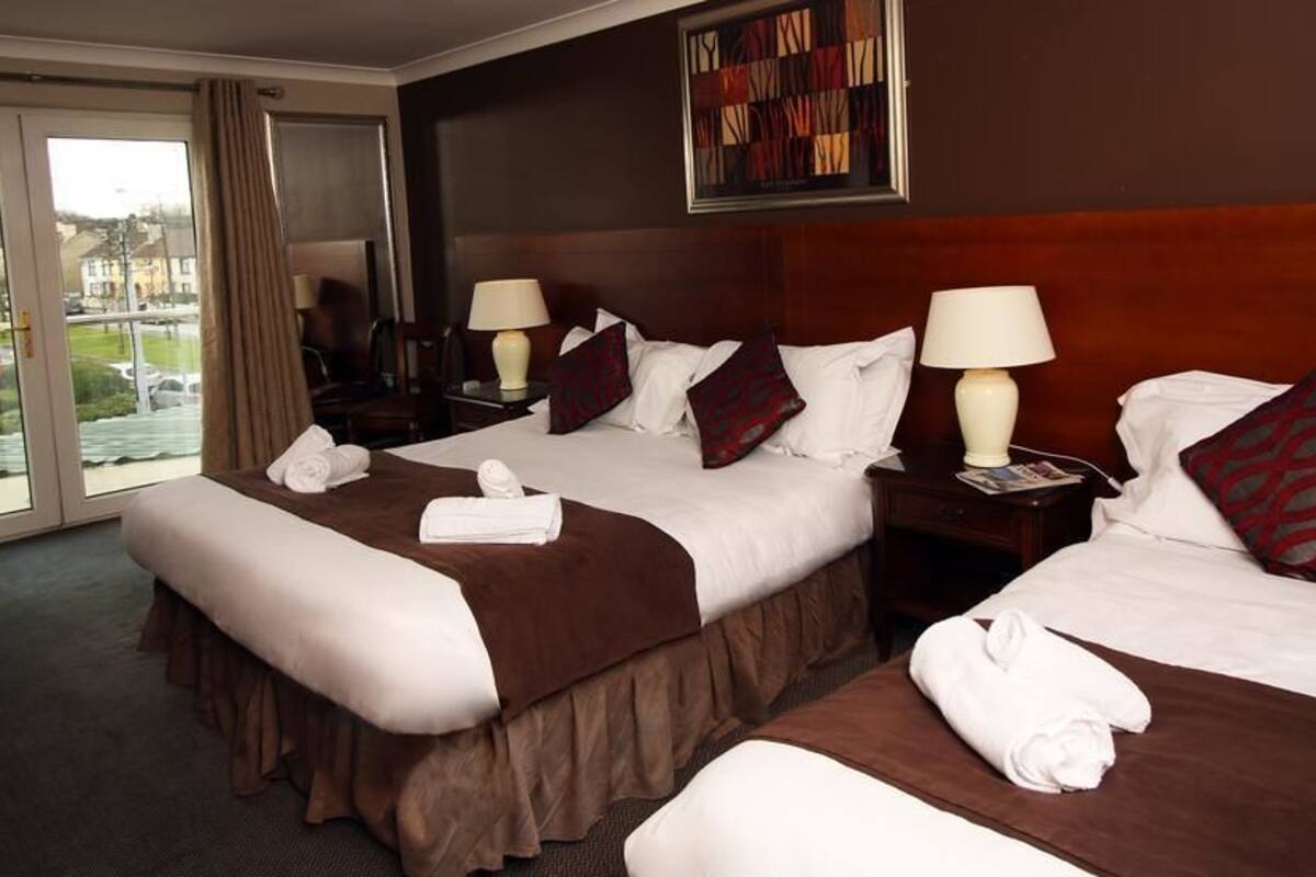 A hotel room with two beds, each with a brown bedspread, white pillows, and a red patterned pillow. There are two bedside tables with lamps, a window with curtains, and a framed artwork on the wall.