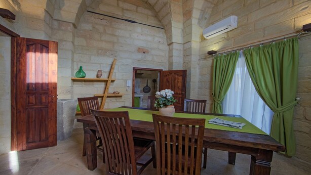 A rustic dining room with a wooden table and chairs, a green tablecloth, and a window with green curtains.