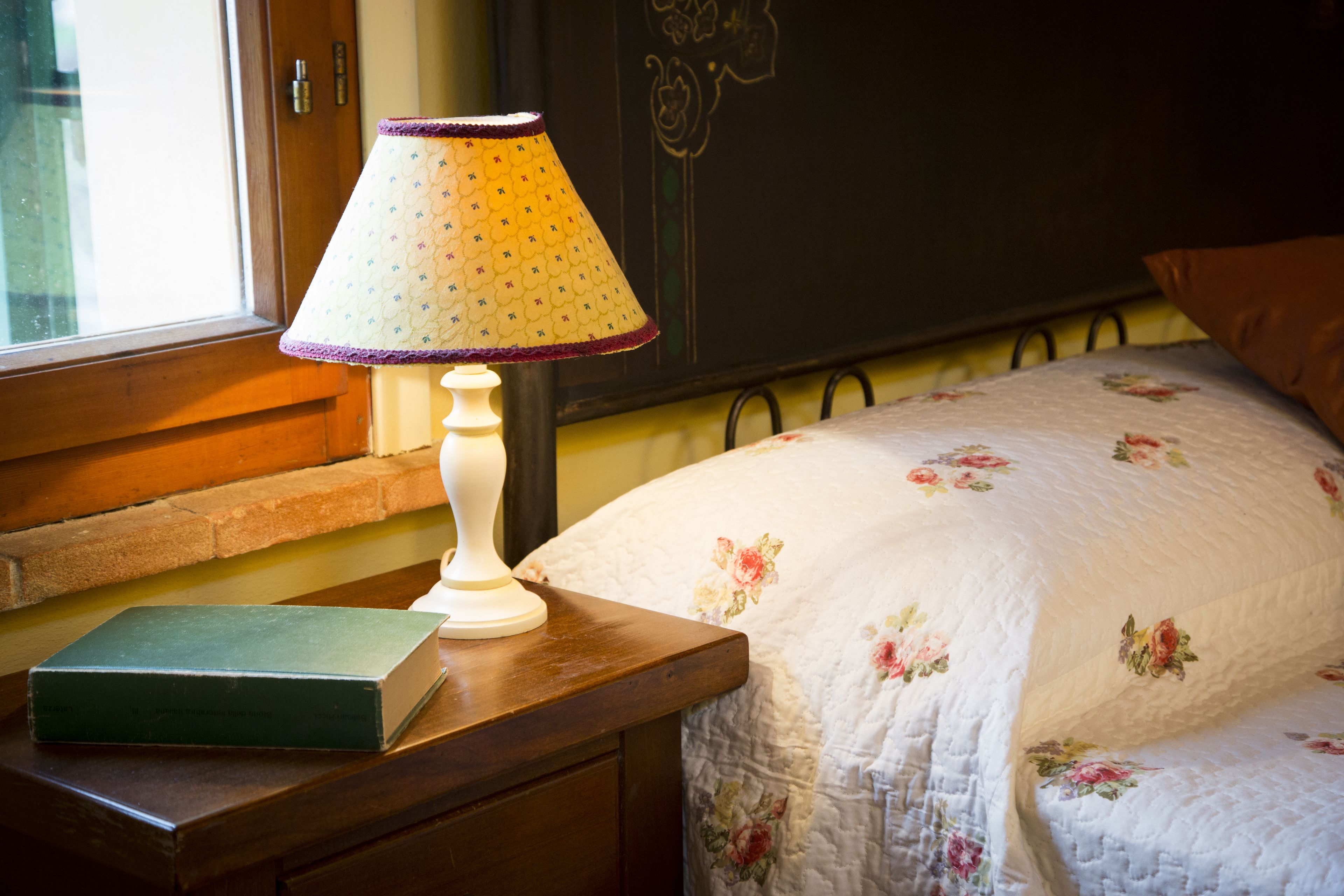 Un lit recouvert d’une couette à motifs floraux, une lampe de chevet et un livre posés sur une table de nuit en bois.