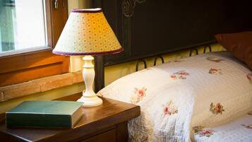 Un lit recouvert d’une couette à motifs floraux, une lampe de chevet et un livre posés sur une table de nuit en bois.