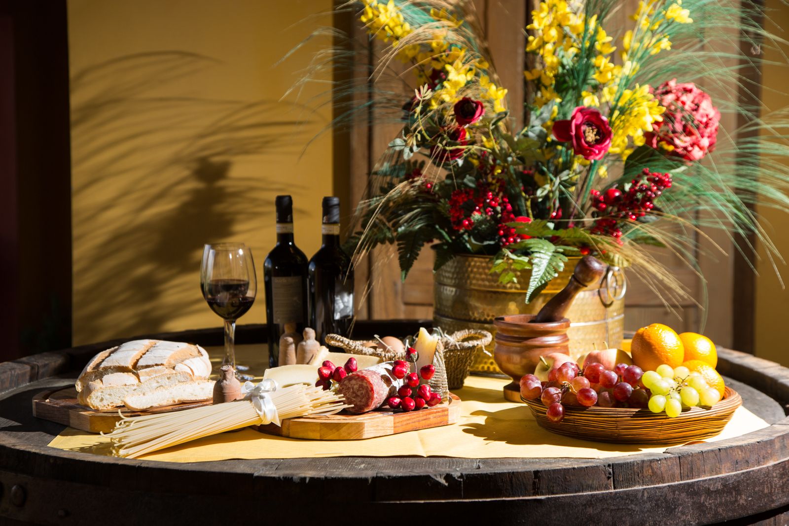 Una mesa redonda de madera con diversos alimentos, como pan, queso, uvas y una cesta de frutas, además de botellas de vino y un arreglo floral.