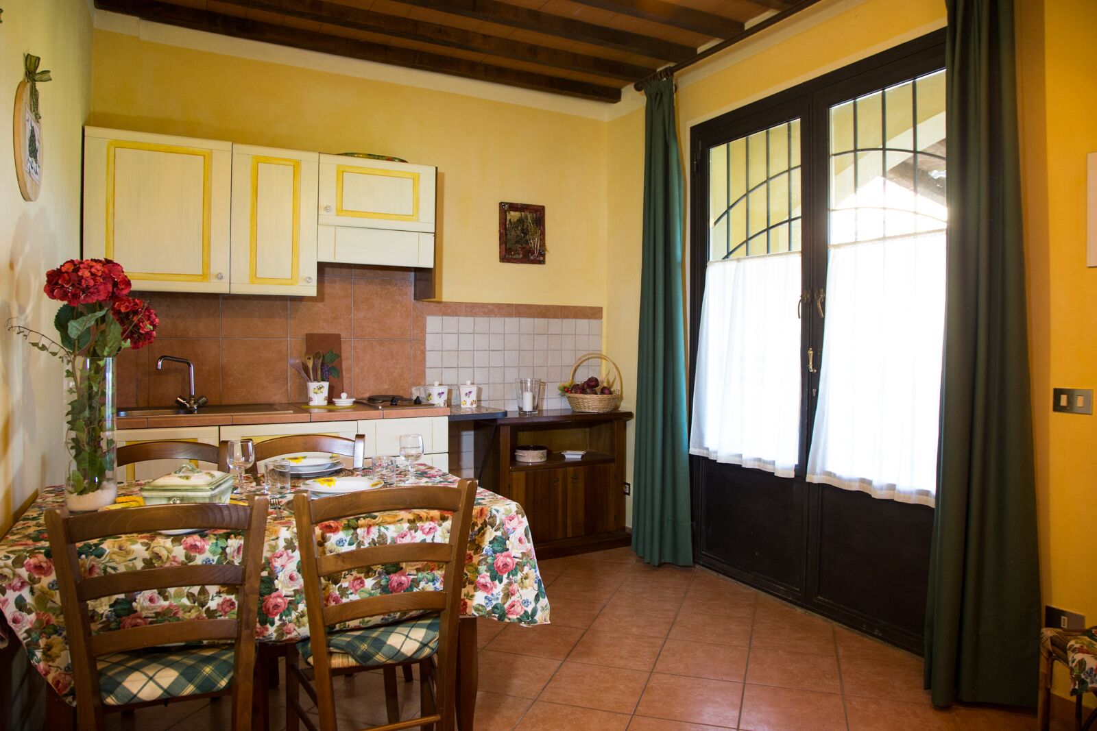 A dining area with a table set for two, a vase of red flowers, and a kitchen in the background.