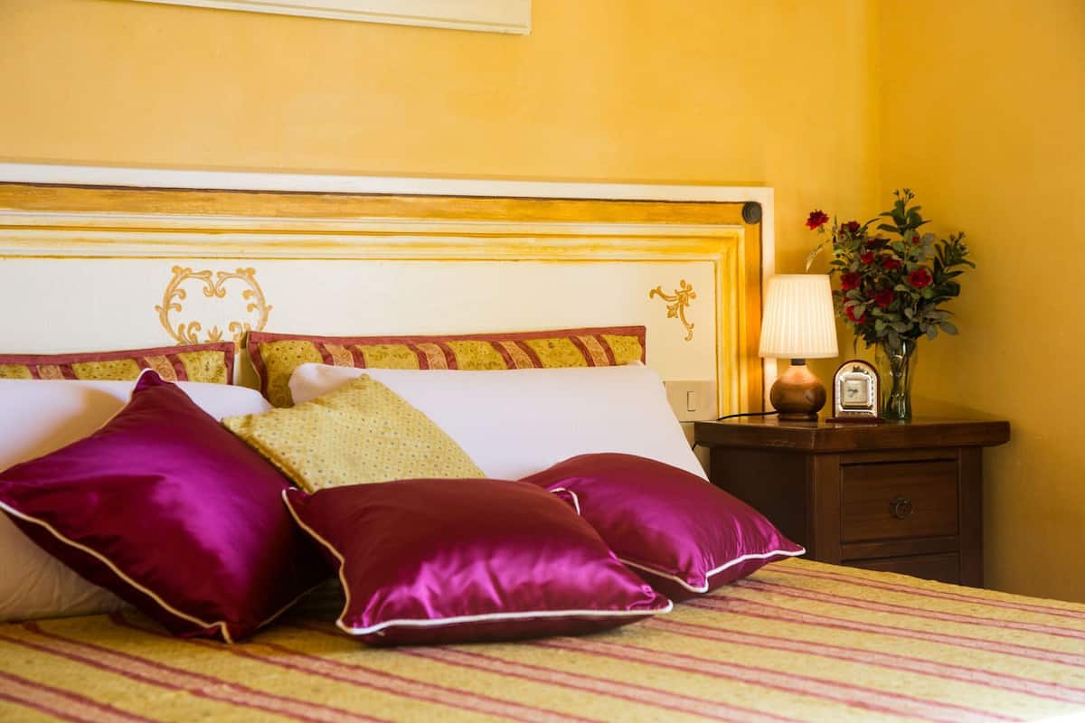 A neatly made bed with a striped bedspread and multiple pillows, a bedside table with a lamp and a clock, and a vase with flowers on the nightstand.
