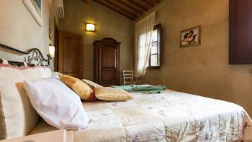 A bedroom with a bed, pillows, a wooden wardrobe, a chair, a window with curtains, and a wall-mounted picture.