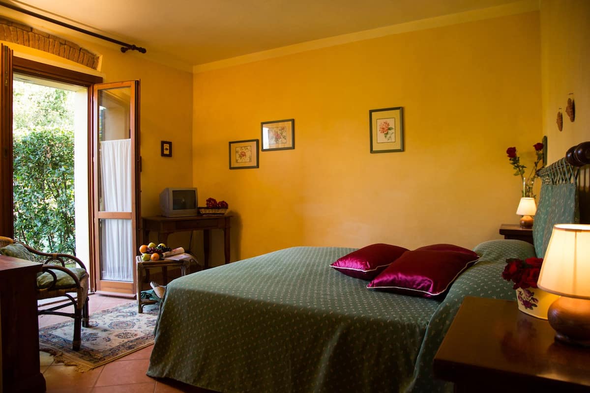 A bedroom with a bed, a desk, a chair, and a window with a view of greenery.