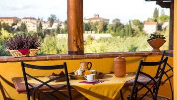 A table set with a yellow tablecloth, two chairs, a basket with purple flowers, a clay pot, and a small potted plant on a balcony with a view of houses and trees.