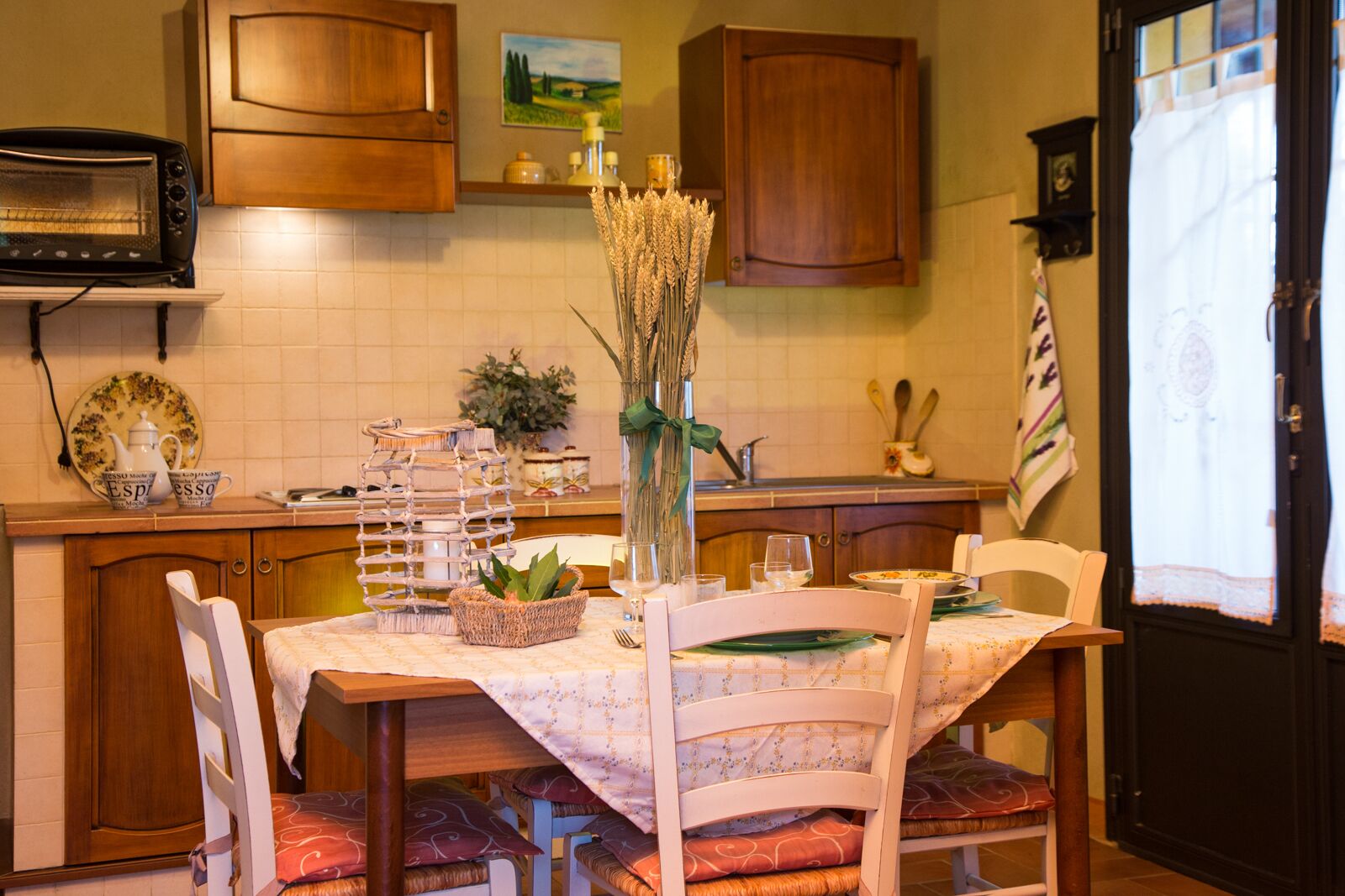 Uma cozinha com armários de madeira, micro-ondas, mesa posta com cadeiras e um vaso com espigas de trigo.