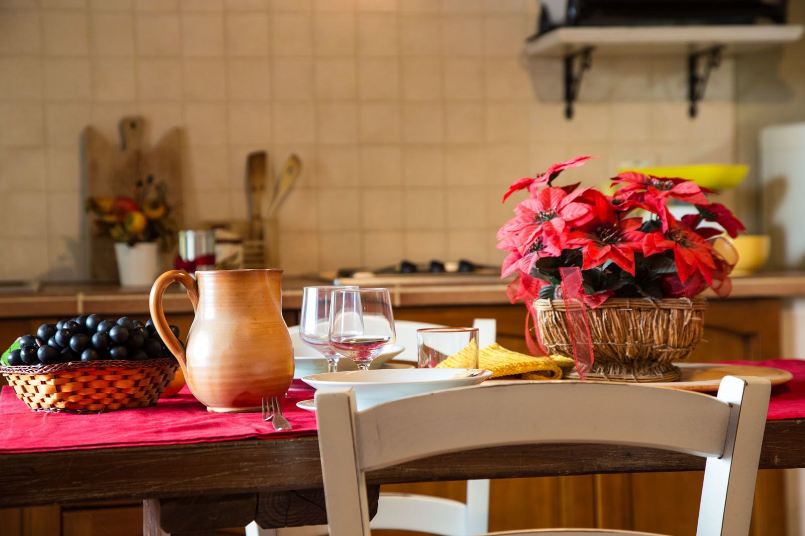 Una cocina con una mesa de madera preparada para comer, una jarra, una cesta de uvas y una planta de estrella.
